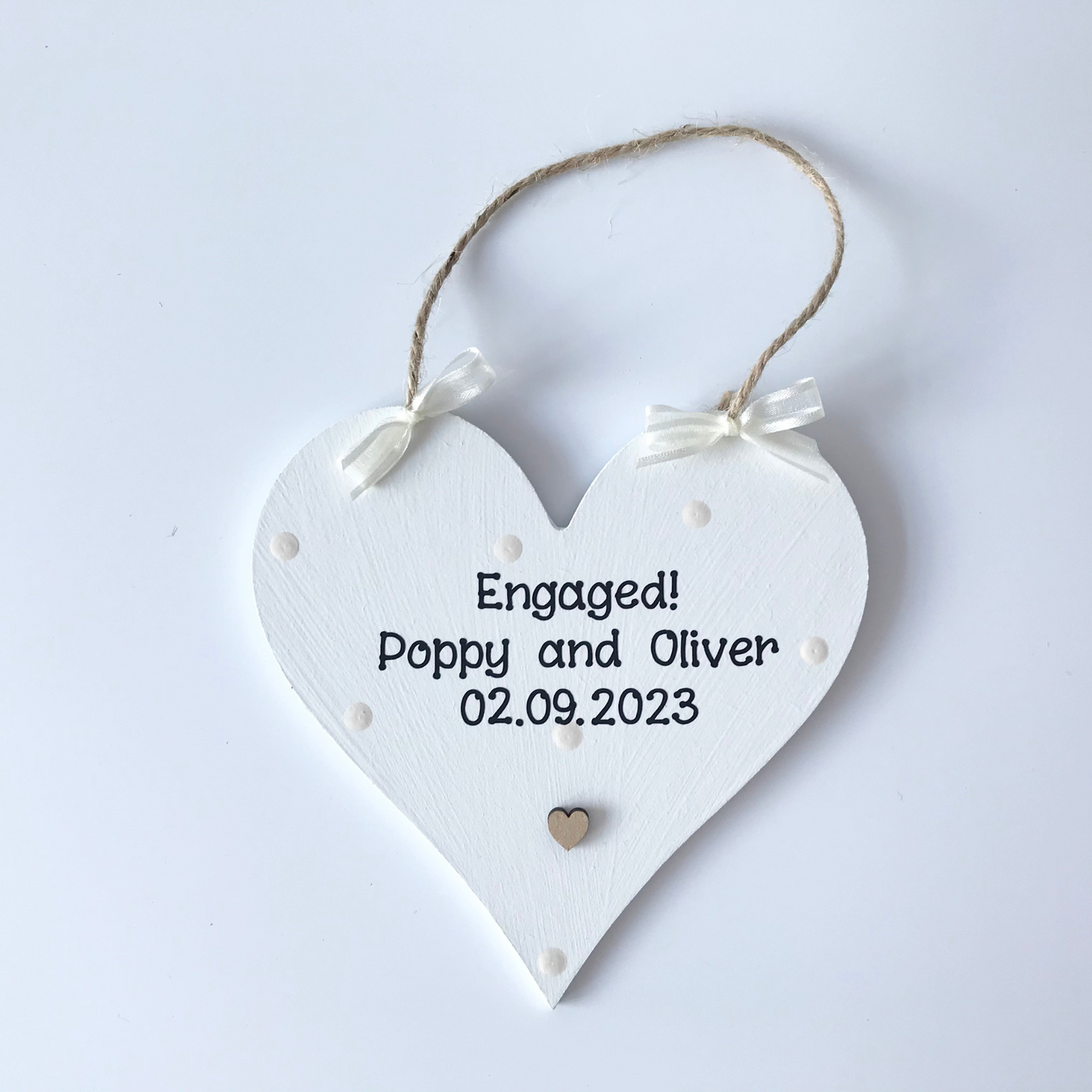 Photo showing white heart plaque with cream dots, cream ribbon, engagement names and date