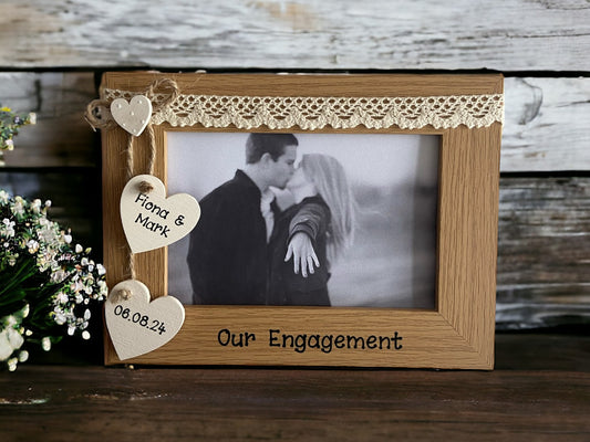 Image shows a brown photo frame for engagements, consists of two cream hanging hearts with names and date of engagement, as well as a small dotted heart to hold together, cream lace runs along the top.