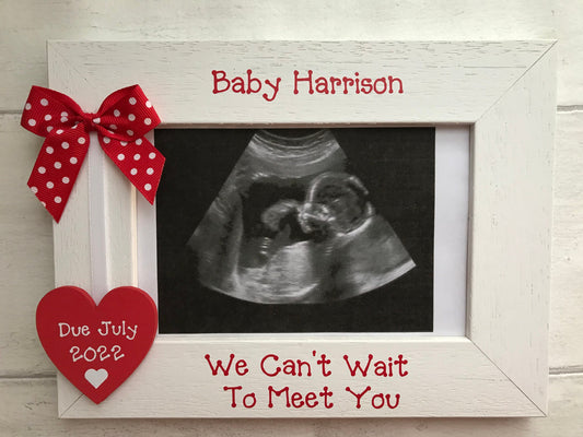 Image shows wooden photo frame for baby scan, consists of wooden heart with due date, attached to white ribbon and a dotted bow.