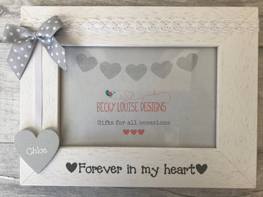 Image shows a wooden photo frame for a lost loved one, consists of a wooden heart with a name attached to white ribbon, dotted bow and white lace along the top.